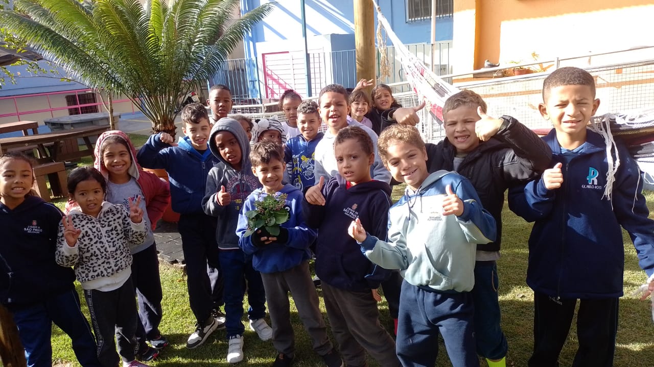 Na imagem, um grande grupo de crianças posam sorrindo para a foto, estão posicionados em uma grama verde, alguns deles fazem sinais de jóia com as mãos e um dos meninos segura um vaso de flores.